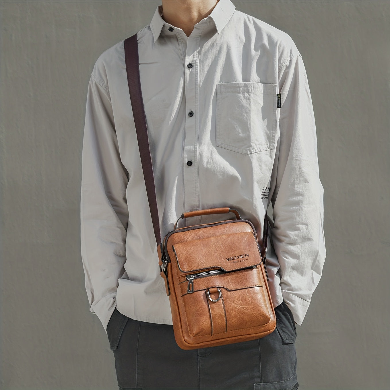 Person wearing a light gray shirt with a brown leather shoulder bag on a gray background