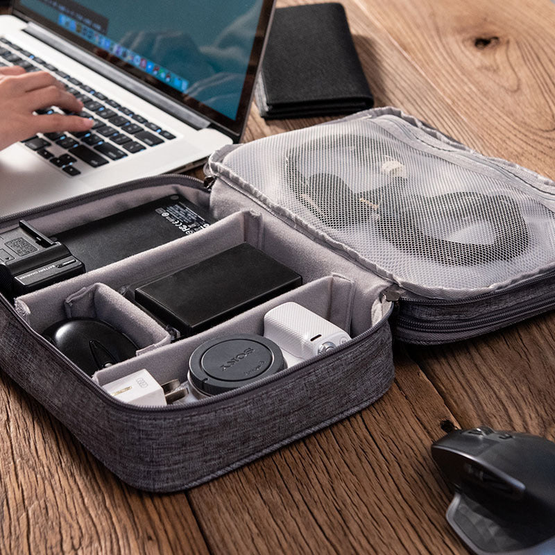 Gray travel case with electronic devices on a wooden desk, person using laptop in the background